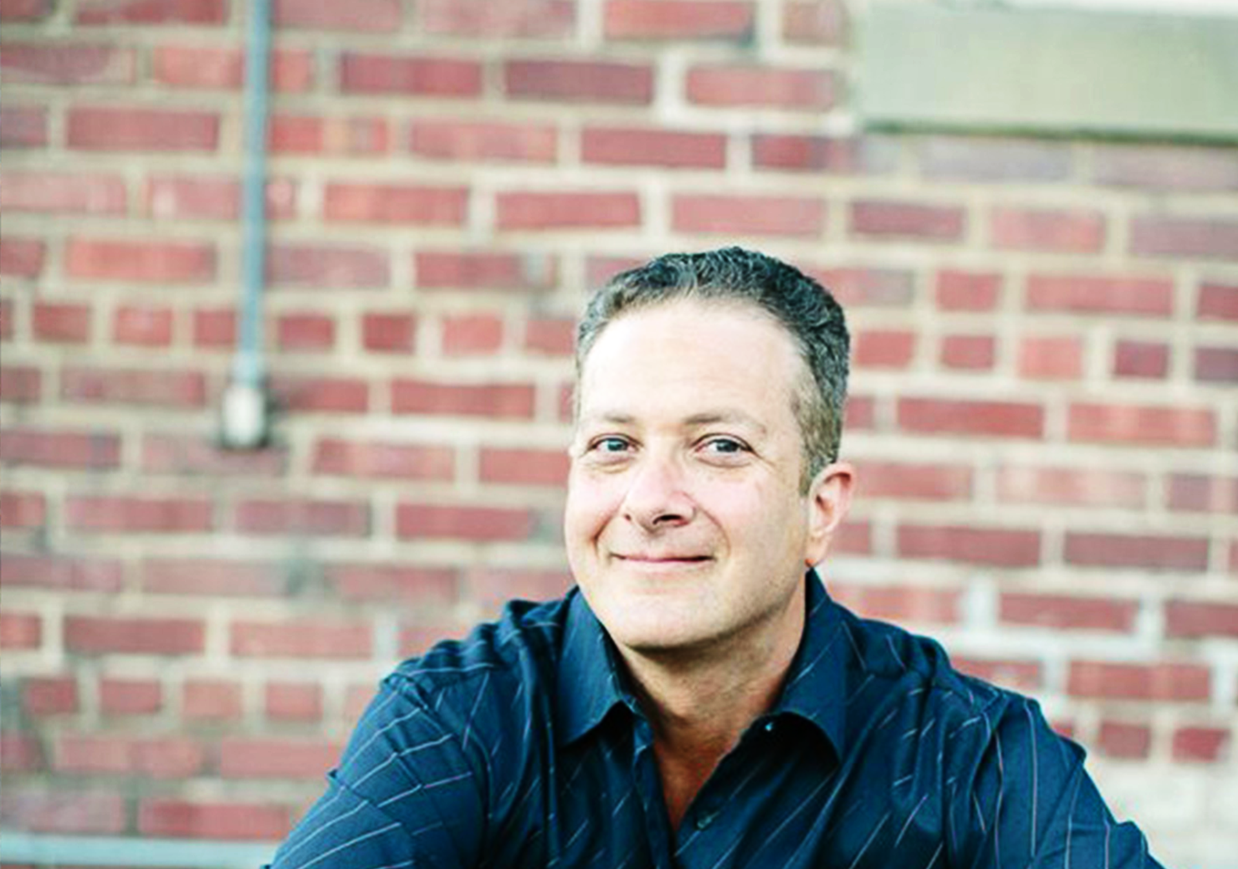 Photo of Peter Baum sitting down, smiling confidently, wearing a navy blue shirt with a brick wall behind him